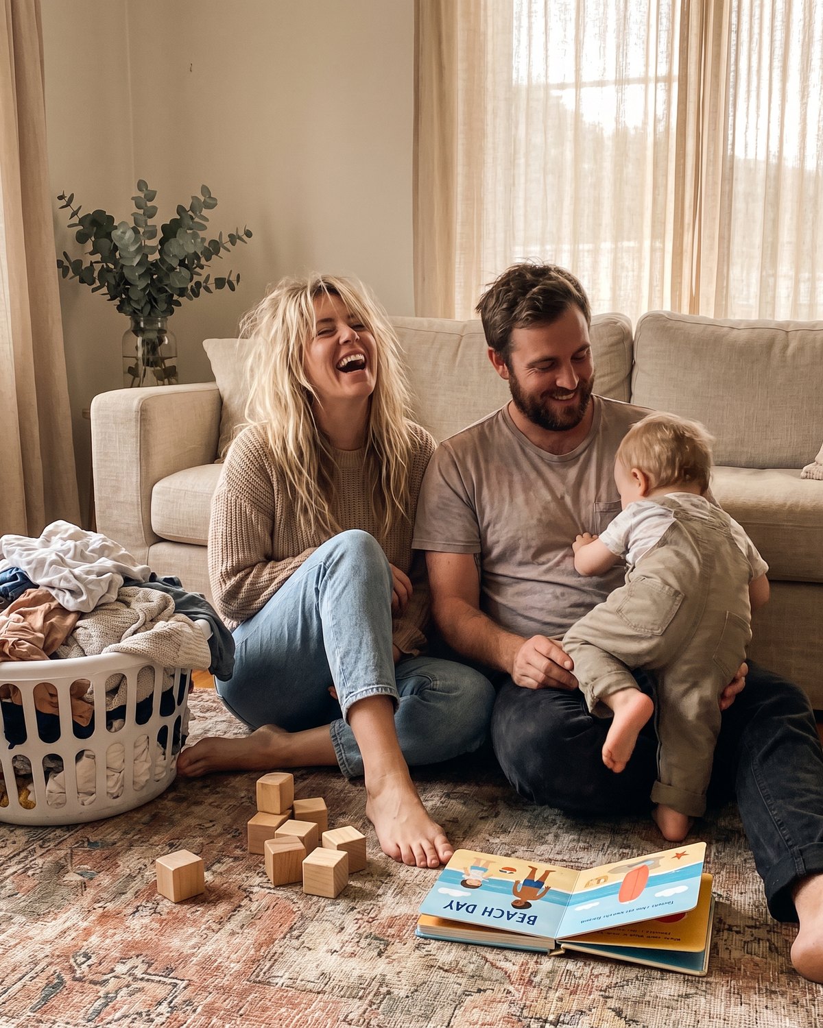 Two parents laughing on the living-room floor with their toddler, washing basket beside them