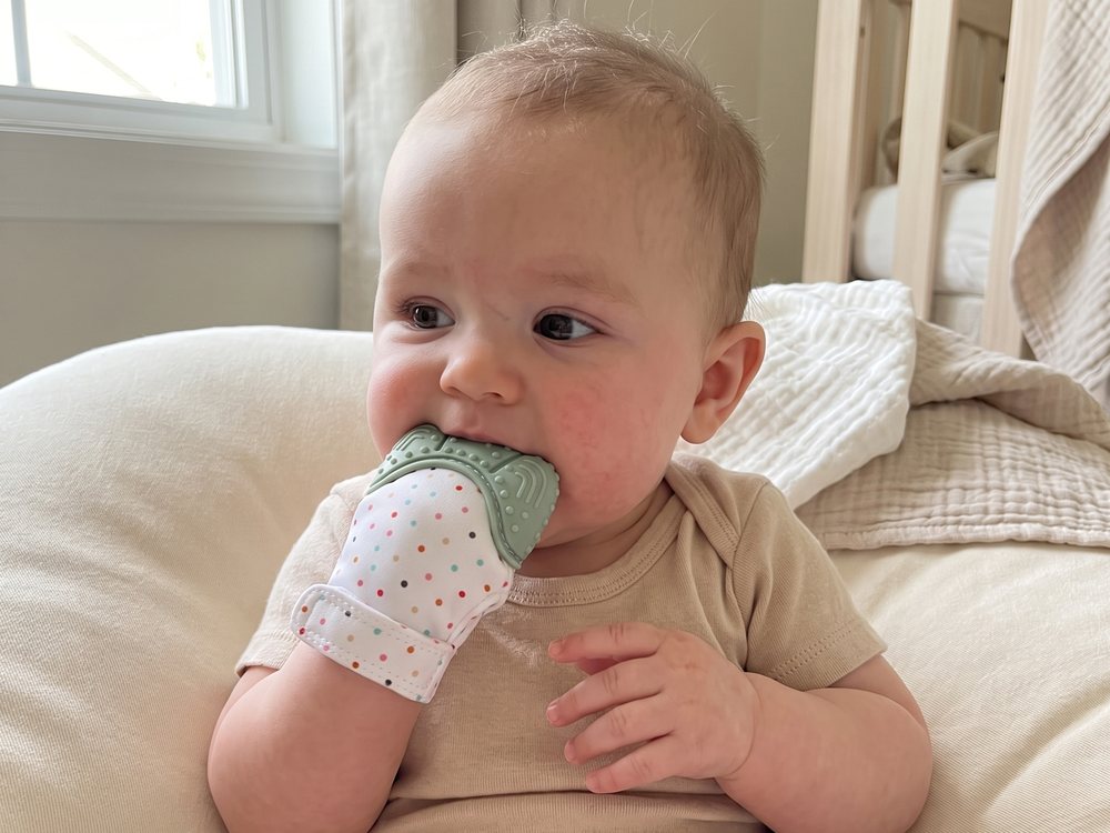 A baby on a cream couch with the mitten in their mouth, other hand free.