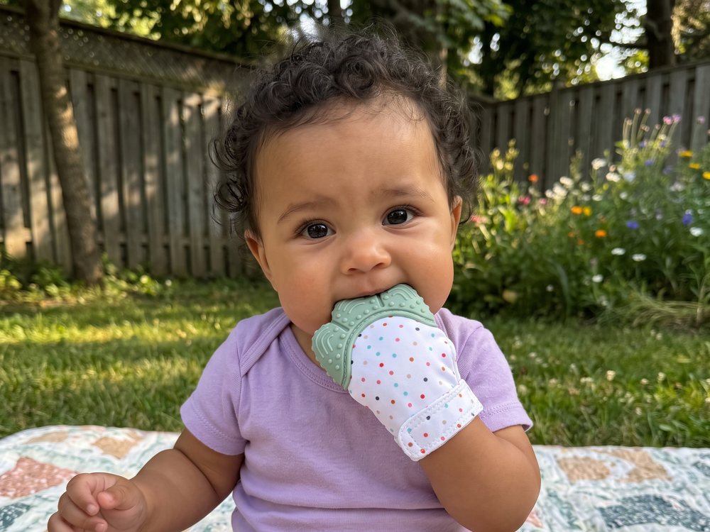A smiling baby in a lilac onesie on a picnic rug in a garden, the mitten on one hand.