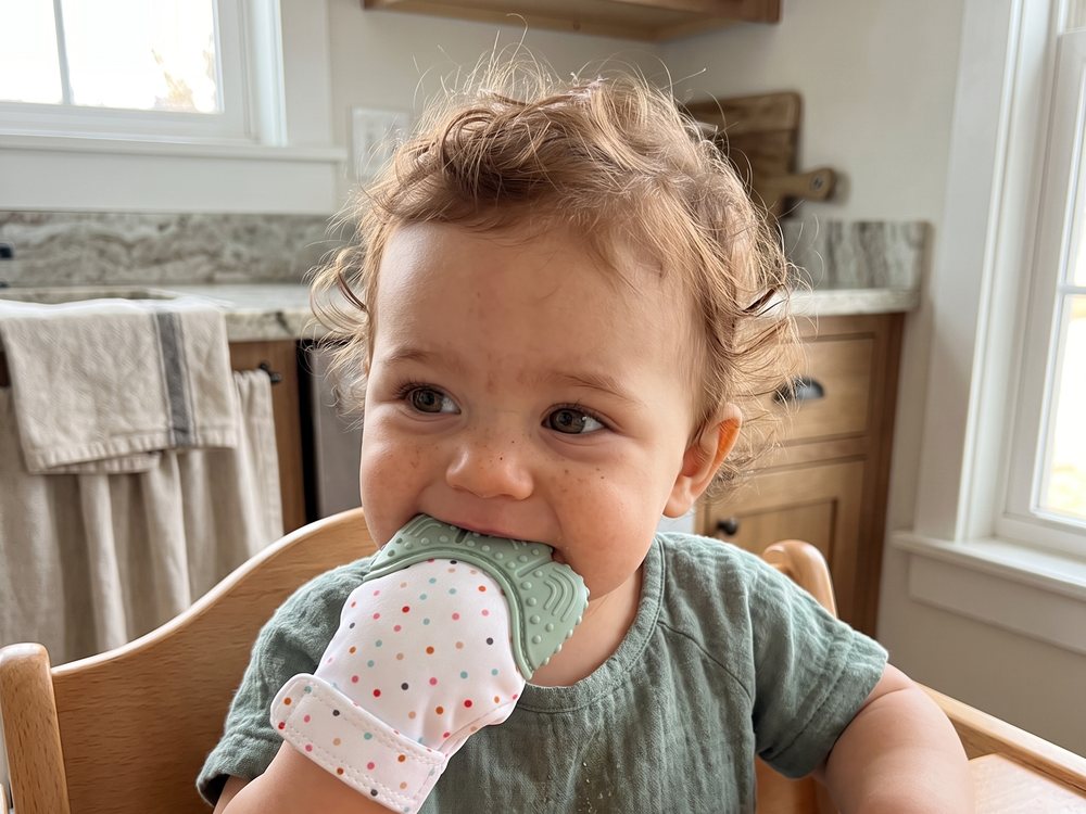 Close view of a toddler in a high chair with the wet mitten in their mouth, silicone panel showing.