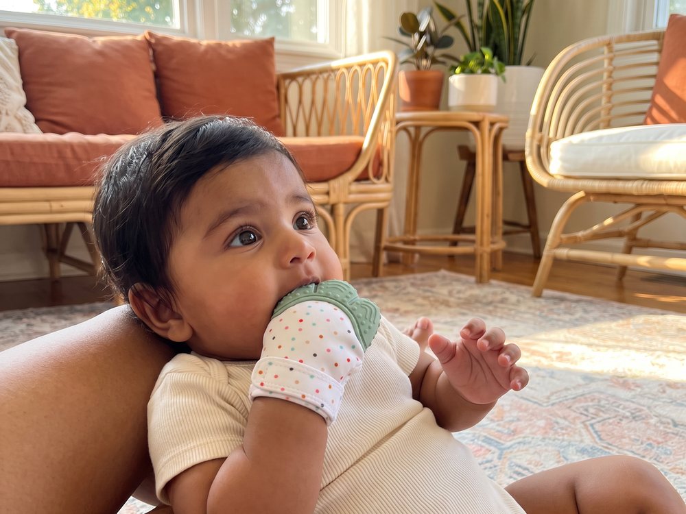 A baby lying across Nanna's lap on the lounge, chewing the silicone panel of the mitten.